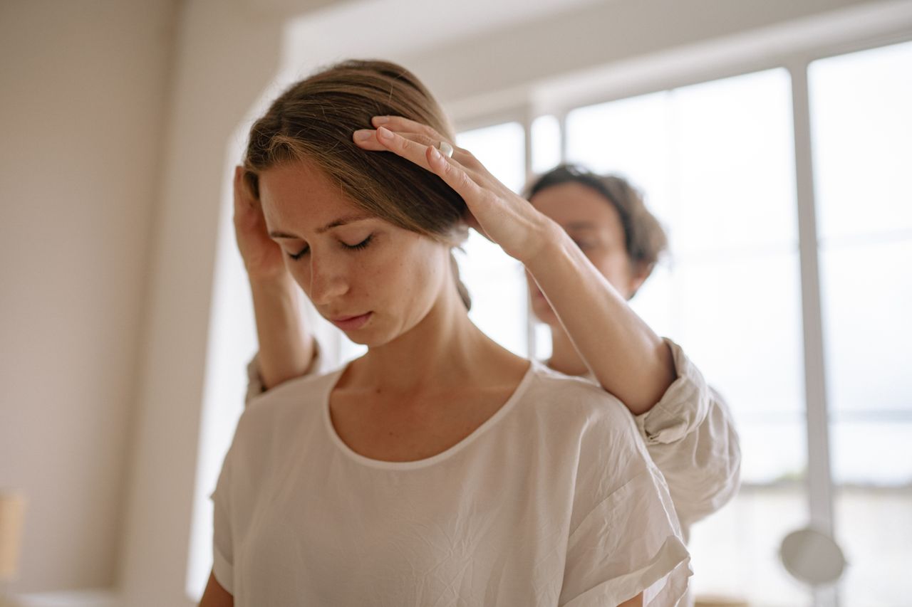 Reiki practitioner tilts a clients head gently forward during a session, backlit by soft, warm window light that feels clean and healing