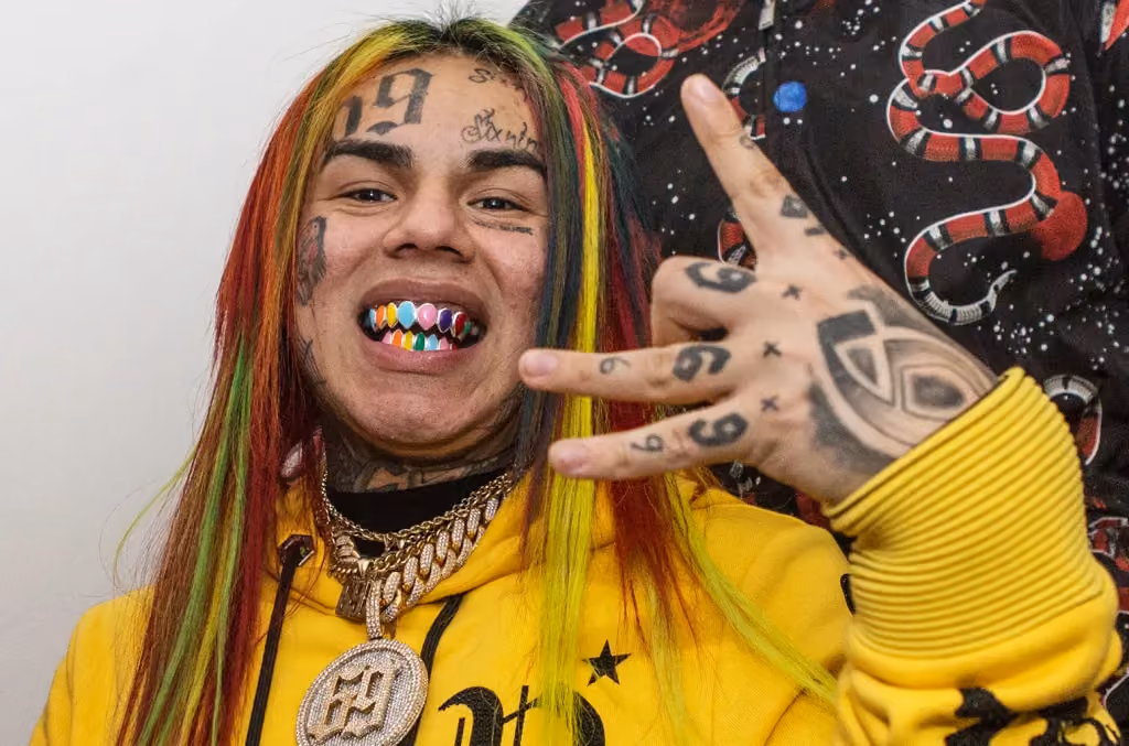 Rapper 6ix9ine poses for the camera in his full artistic getup featuring bling, rainbow hair, rainbow grill, and of course face and hand tattoos