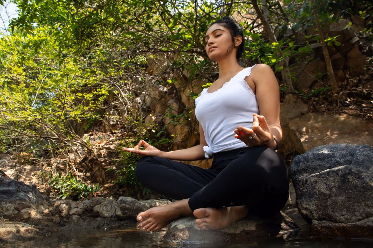 woman sitting in lotus position at the edge of a creek, meditating on the rocks to take advantage of the spiritual power and meaning of water