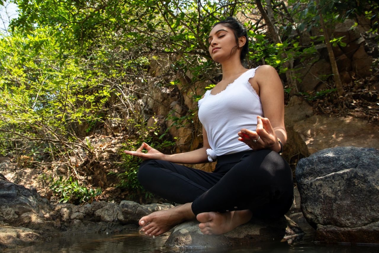 woman sitting in lotus position at the edge of a creek, meditating on the rocks to take advantage of the spiritual power and meaning of water