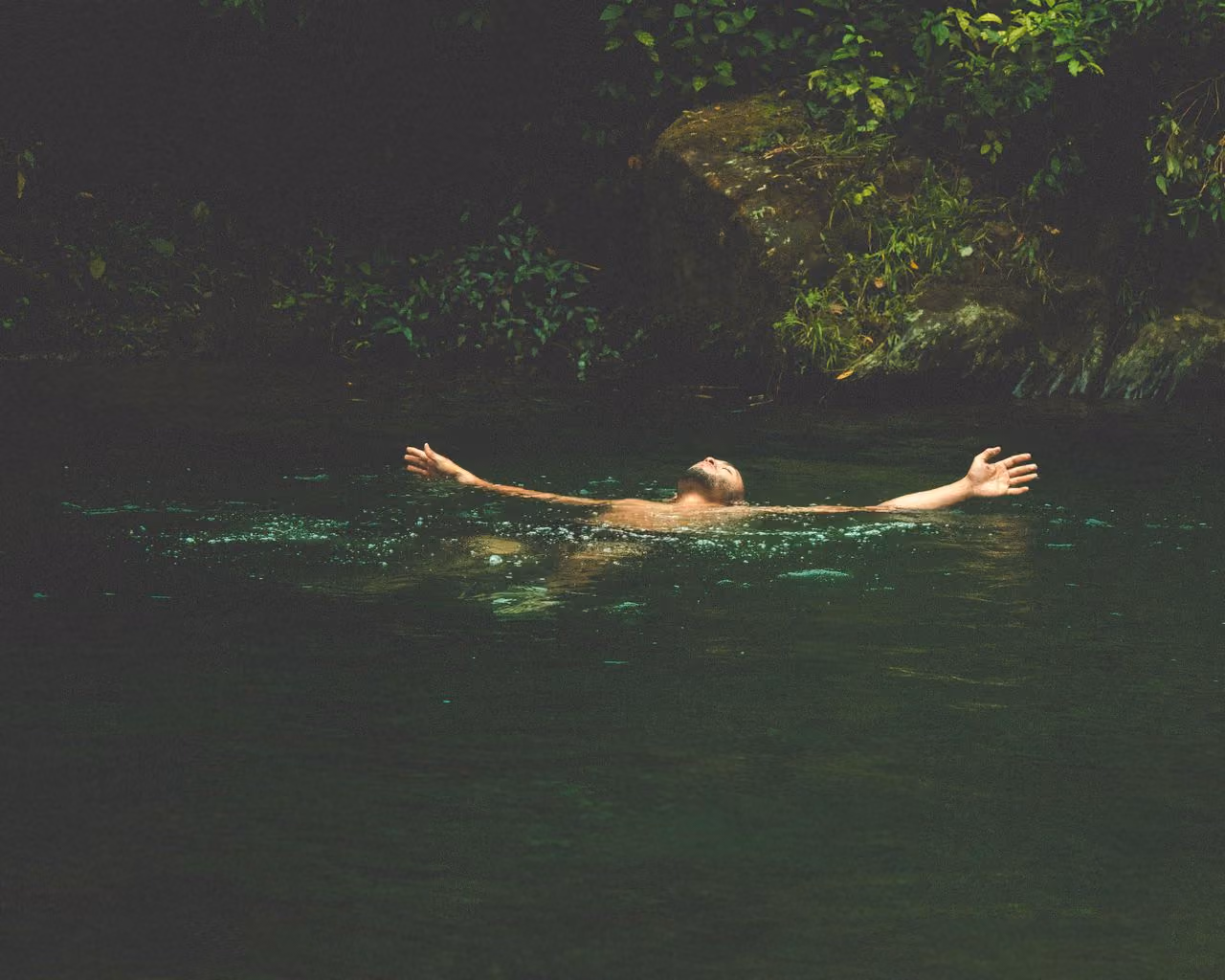 man floating on his back through a natural pool surrounded by lush foliage, arms outstretched in a position of spiritual peace