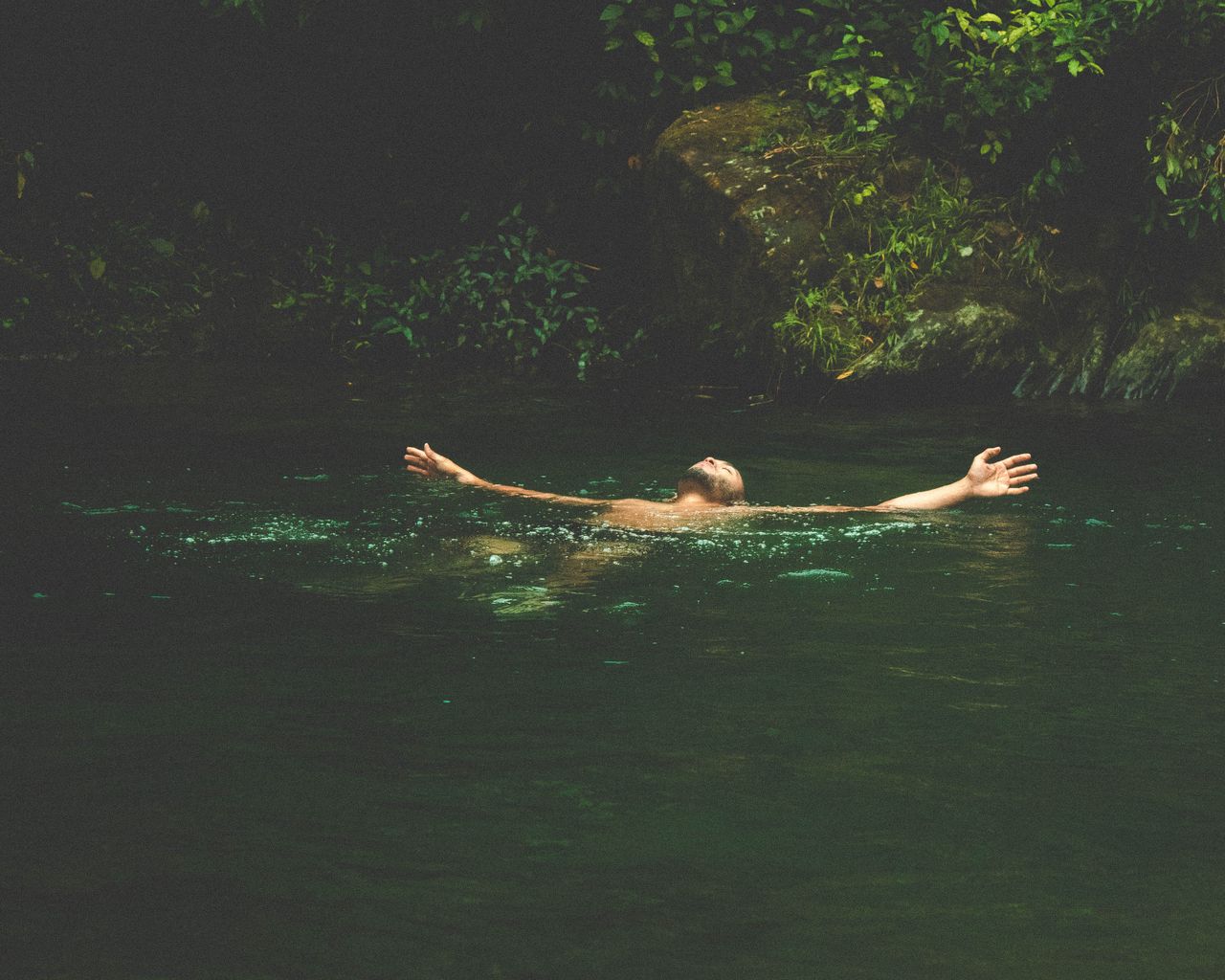 man floating on his back through a natural pool surrounded by lush foliage, arms outstretched in a position of spiritual peace