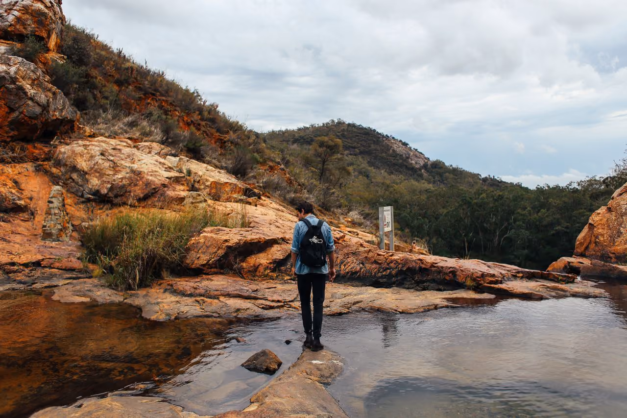 young man in a backpack standing at the edge of a river that cuts through a rough red rock hillside