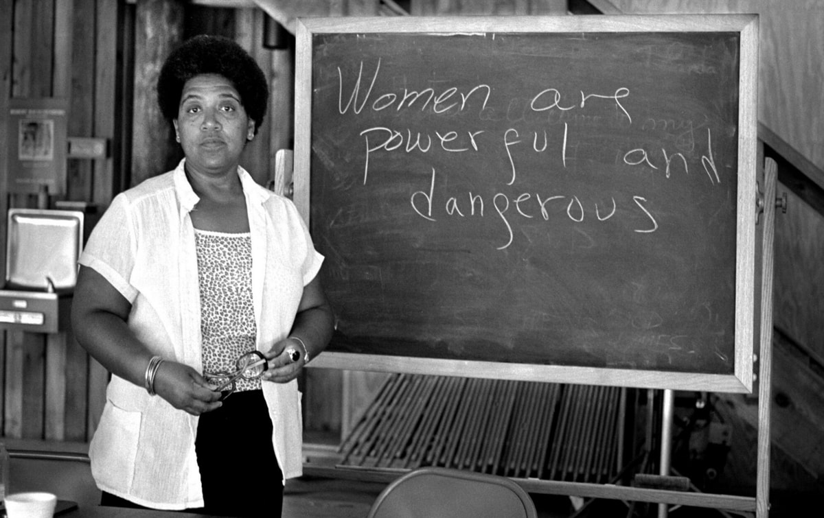 Audre Lorde standing beside a chalkboard bearing the message "Women are powerful and dangerous"
