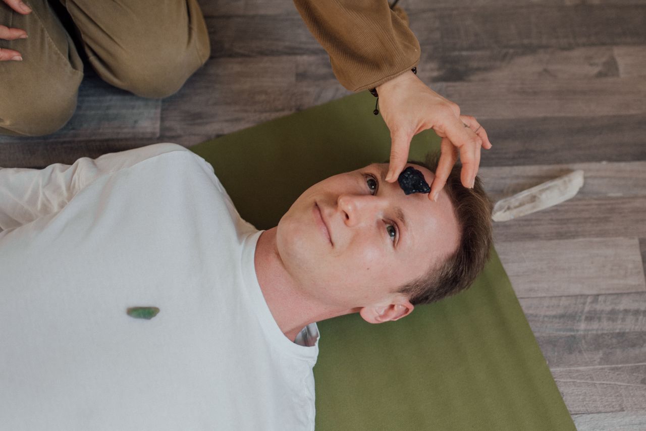 man lying back on a yoga mat as a spiritual practioner places lapis lazuli on his forehead over the third eye chakra