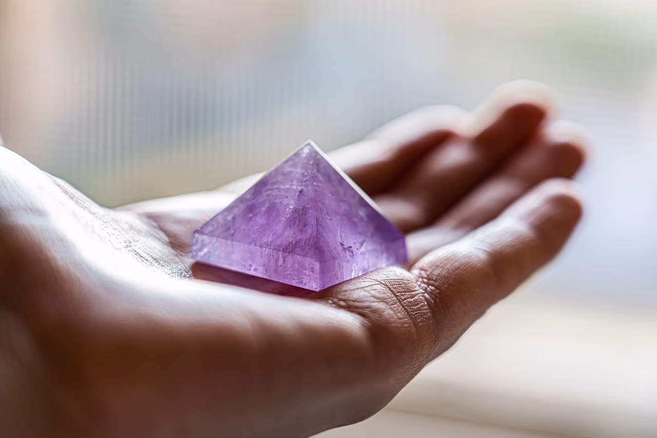 person holding a small pyramid made of amethyst in their balm so that it catches the light, highlighting the swirls of color contained within the gemstone's purple body
