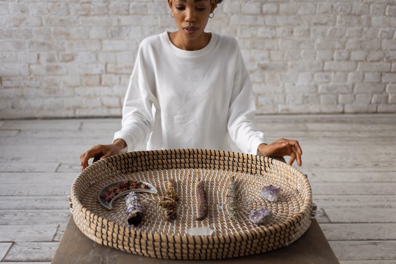 woman seated before a basket with tools for cleansing a spiritual space, including amethyst and sage smudge sticks