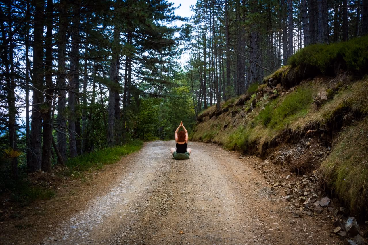 woman seated on the earth on a forest trail, hands pressed together overhead in a pose of meditative spiritual reflection