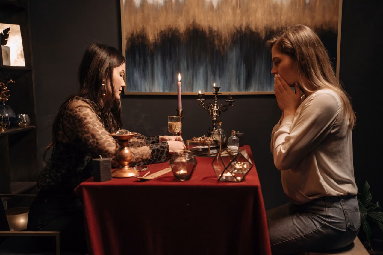 two women across the table from one another during a spiritual reading; one, a shop owner peddling services and crystals, the other, a young woman looking shocked, trying to decide what fits into her spirituality and what does not