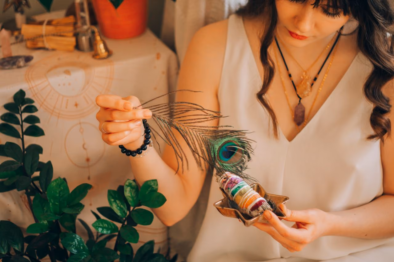 A woman holding a smoldering sage smudge bundle and wafting it with a peacock feather — representing the practice of smudging for energy clearing and space cleansing.