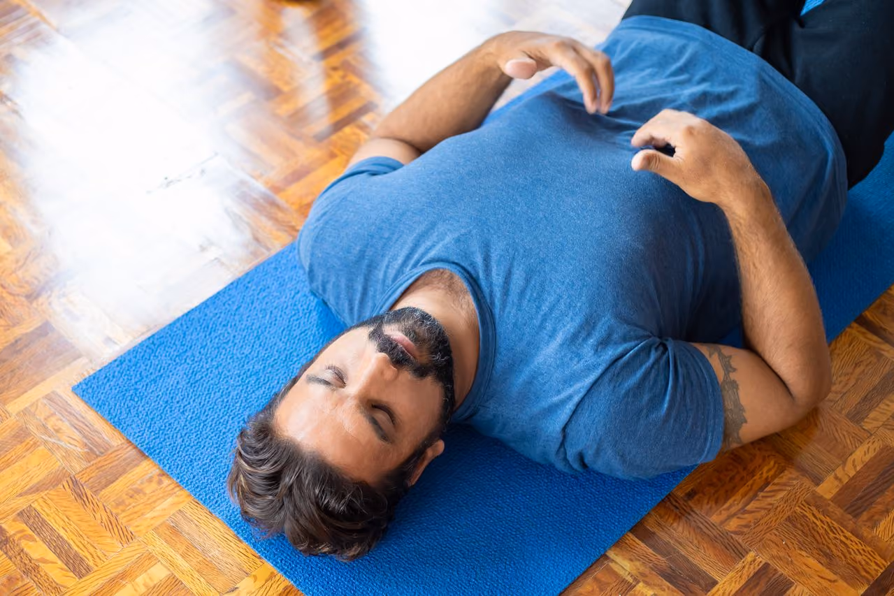 man lying on his back on a yoga mat, placing his fingers below the ribs to monitor breath as he meditates