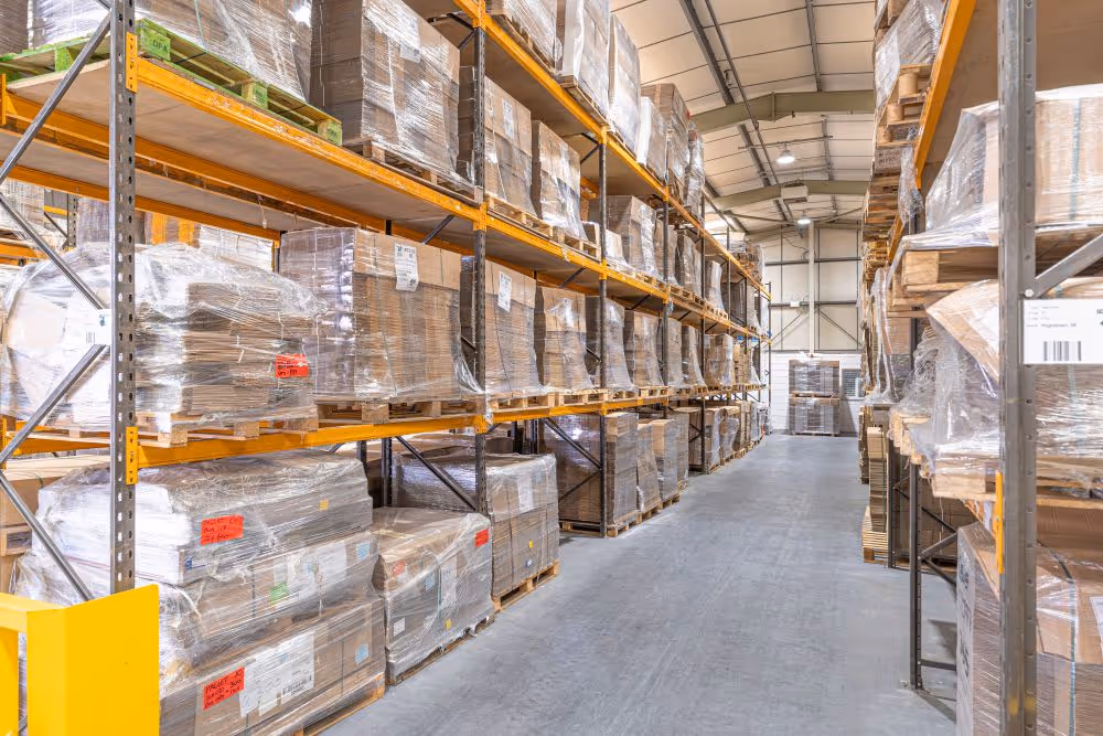 Warehouse aisle with tall metal racks holding stacked pallets wrapped in plastic.