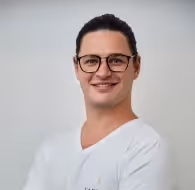 Smiling man with glasses wearing a white shirt against a plain background.