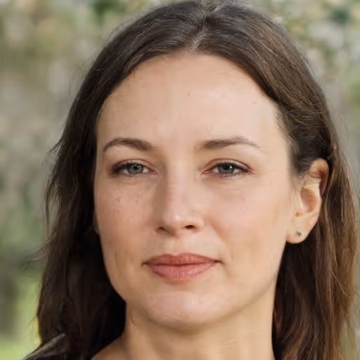Close-up portrait of a woman with light skin, brown hair, and green eyes, with a neutral expression and blurred outdoor background.