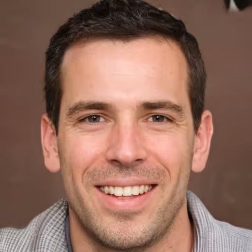 Smiling man with short dark hair and light stubble wearing a gray sweater against a brown background.