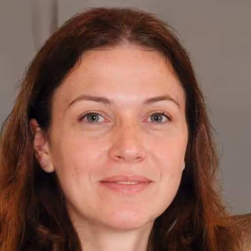 Close-up portrait of a woman with light skin, brown hair, and green eyes smiling gently against a gray background.