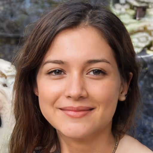 Close-up portrait of a smiling woman with brown hair and brown eyes against a blurred outdoor background.