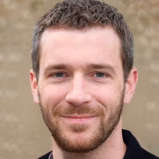 Close-up of a smiling man with short brown hair and beard, wearing a black shirt, against a blurred beige background.