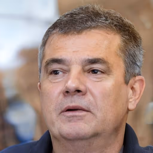 Close-up of a middle-aged man with short gray hair wearing a dark shirt, looking slightly to the side.