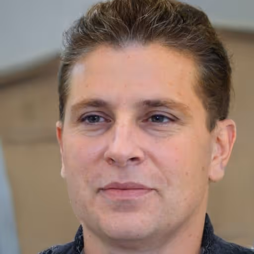 Portrait of a man with short brown hair and blue eyes, wearing a dark top, against a blurred indoor background.