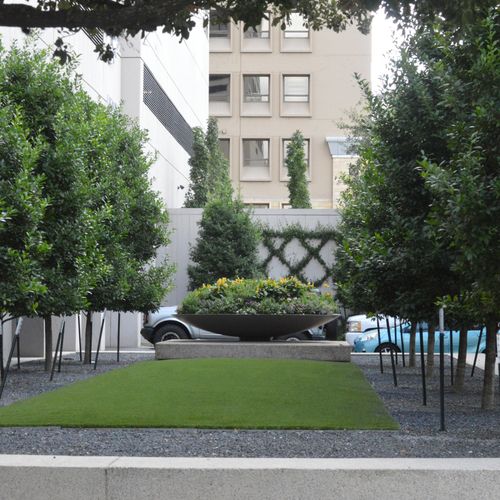 Urban garden with a green lawn, symmetrical rows of trees, and a round concrete planter filled with yellow flowers in front of a building.