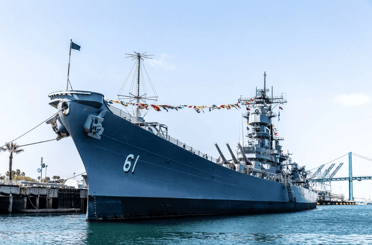 USS Iowa battleship docked at a waterfront with flags and harbor cranes