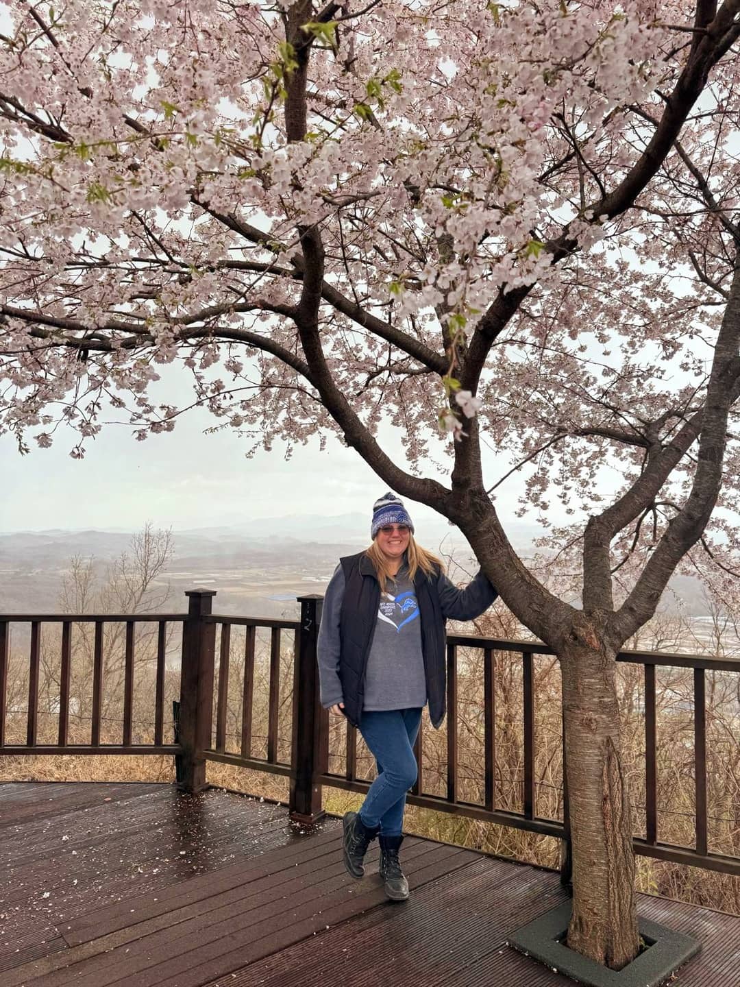 A Woman standing besides the cherry blossom tree and smiling for a picture.