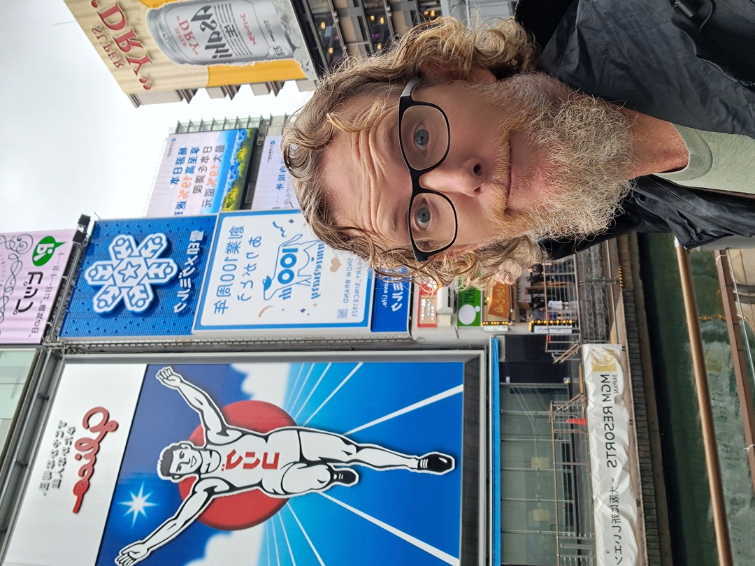 A man wearing glasses stands in front of a large billboard displaying an advertisement of a running man.