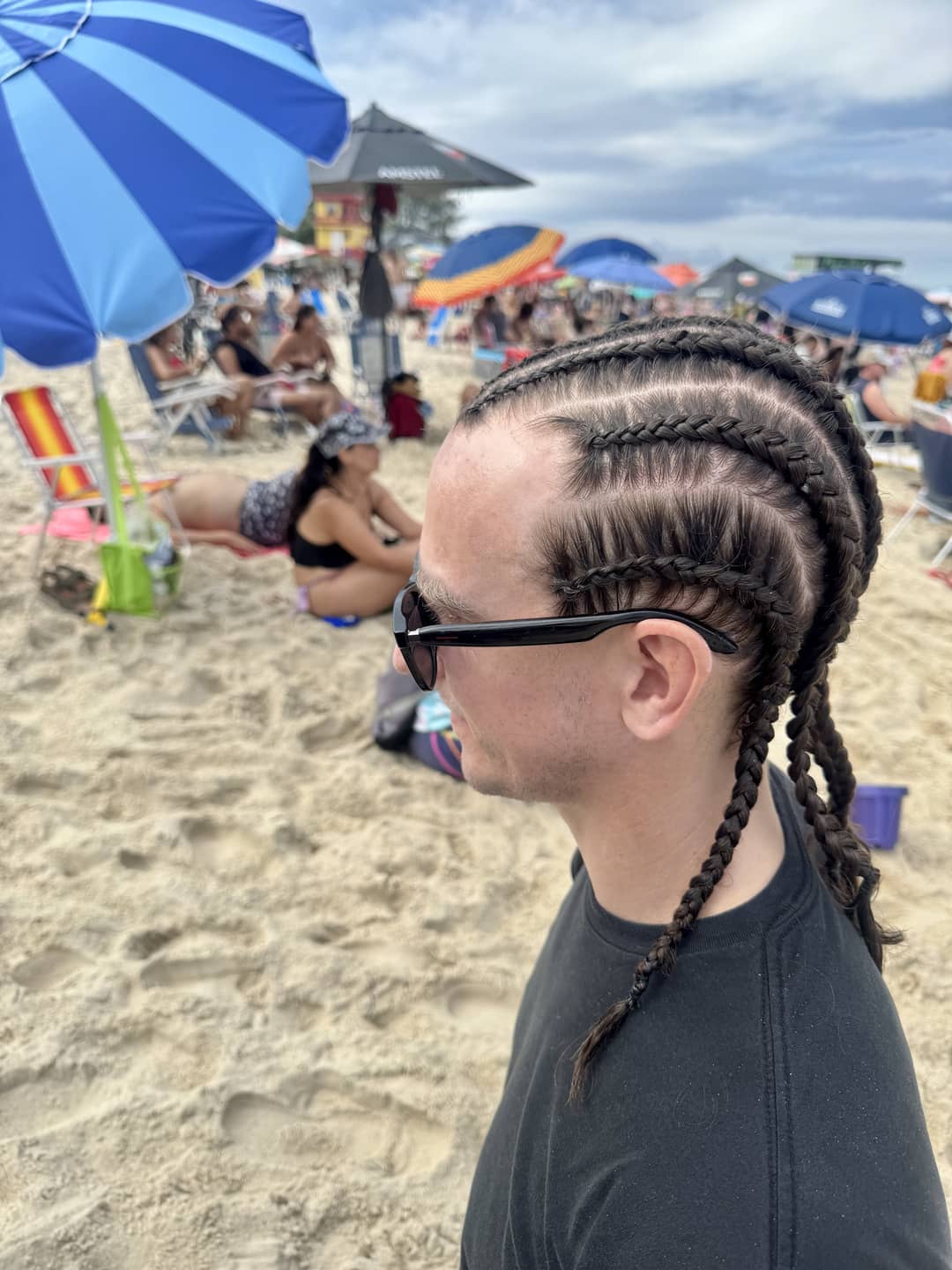A person with braided cornrows and sunglasses standing on a crowded sandy beach with umbrellas and people relaxing in the background.