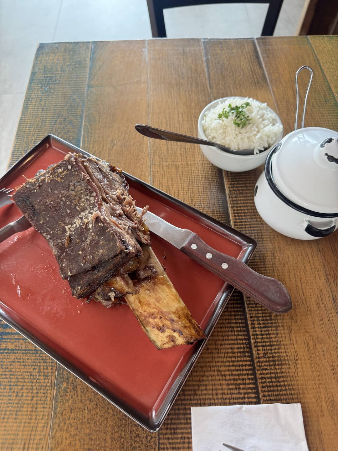 A large bone-in cut of slow-cooked beef served on a red tray with a knife, accompanied by white rice and a small pot on a wooden table.