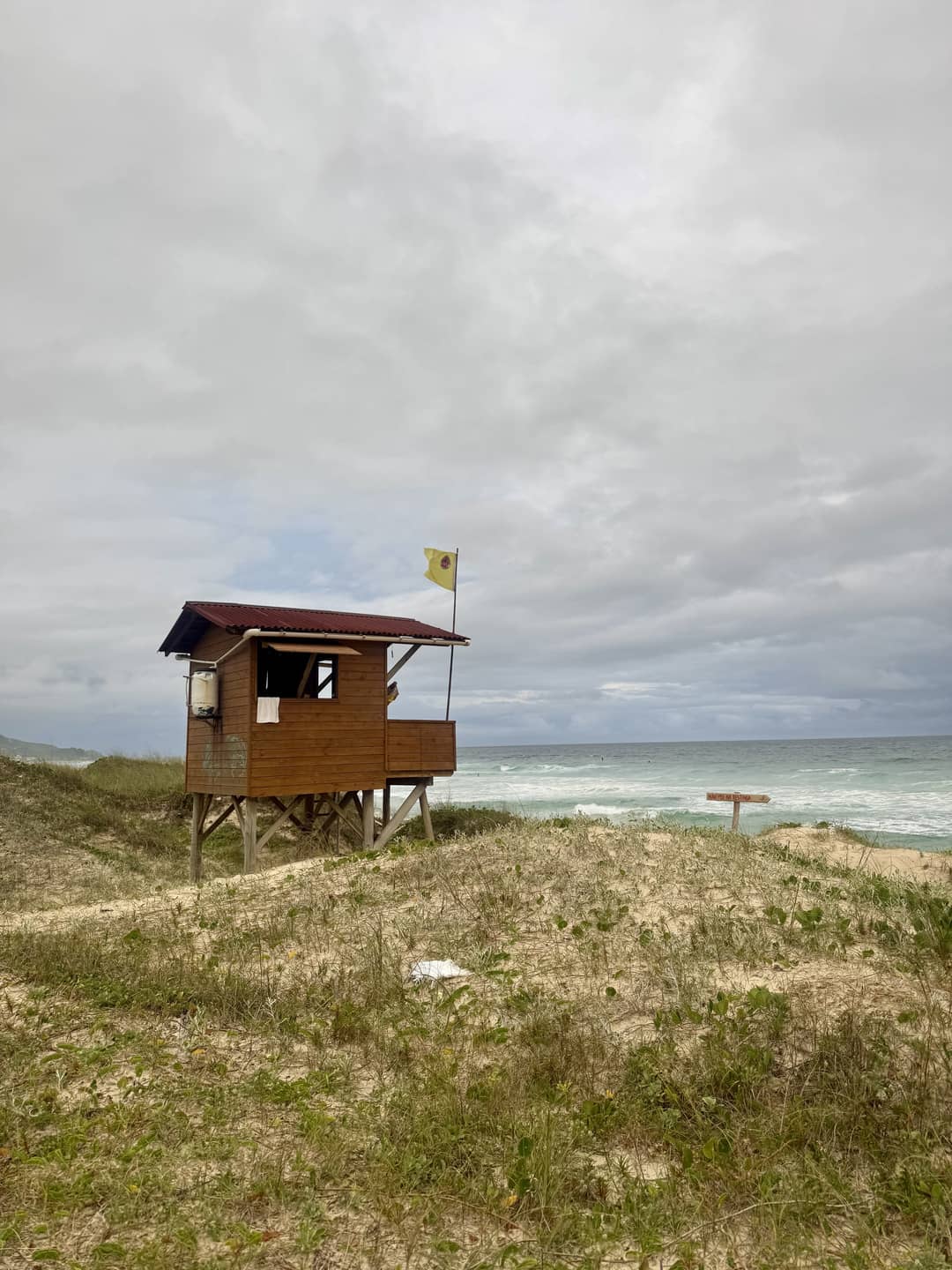 A wooden lifeguard hut on stilts overlooking a sandy beach and ocean, with a yellow flag flying under an overcast sky.