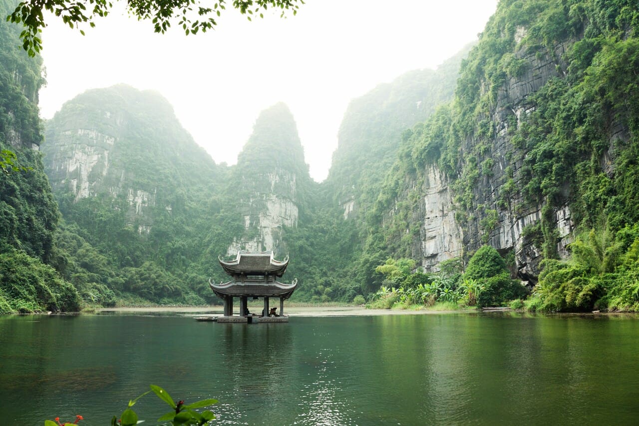 Trang An Scenic Landscape Complex in Ninh Binh, Vietnam, featuring a traditional pavilion surrounded by limestone karst mountains and emerald waters.