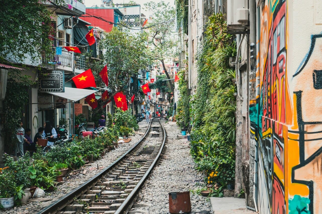 Hanoi Train Street in Vietnam lined with colorful buildings, Vietnamese flags, street art, and railway tracks through the Old Quarter.