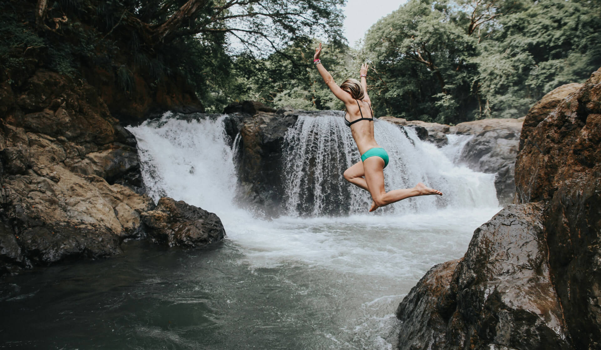 A woman in a bikini jumping off a rock into a pool of water with a waterfall behind her in Costa Rica