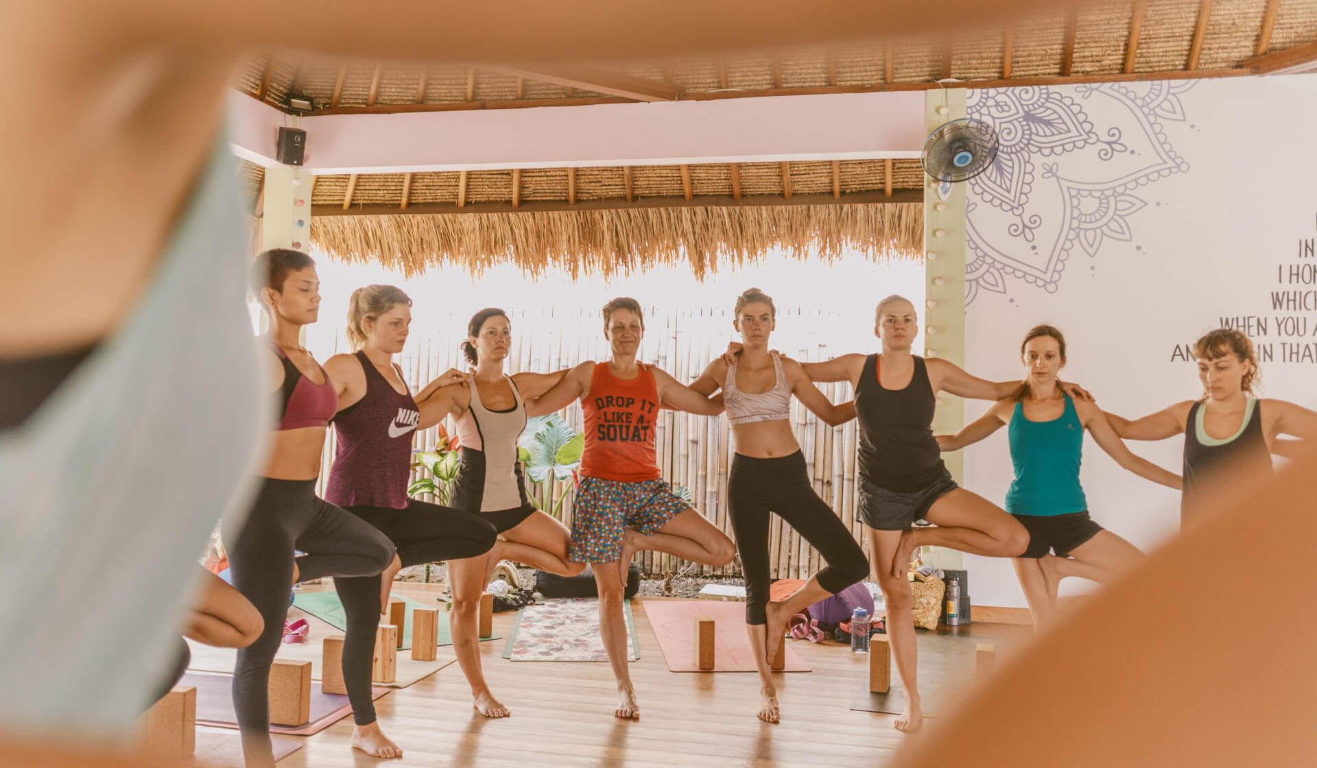 A class of yoga students practicing tree pose in a circle with their arms around each other at a Nomad Yoga 200 hour yoga teacher training