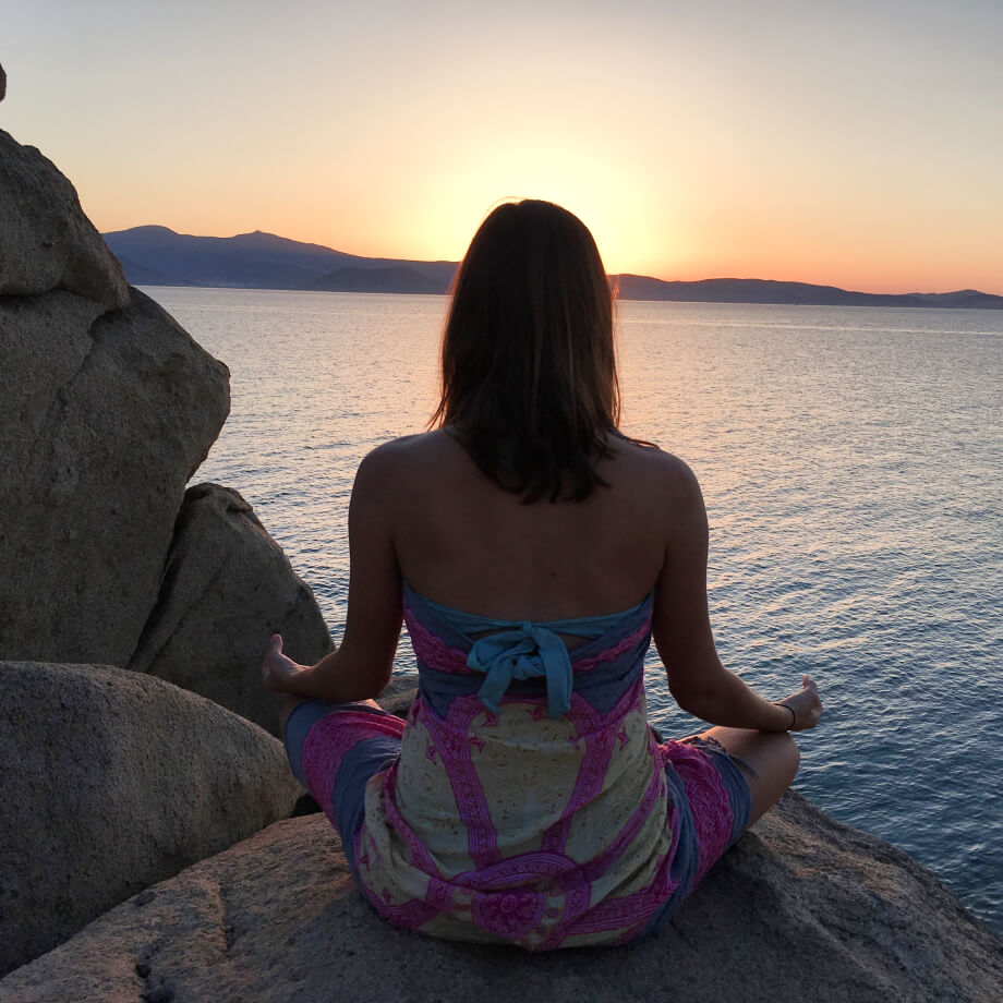 A back view of a young women seated in meditation on gray rocks watching a colorful sunset in front of crystal clear blue sea waters