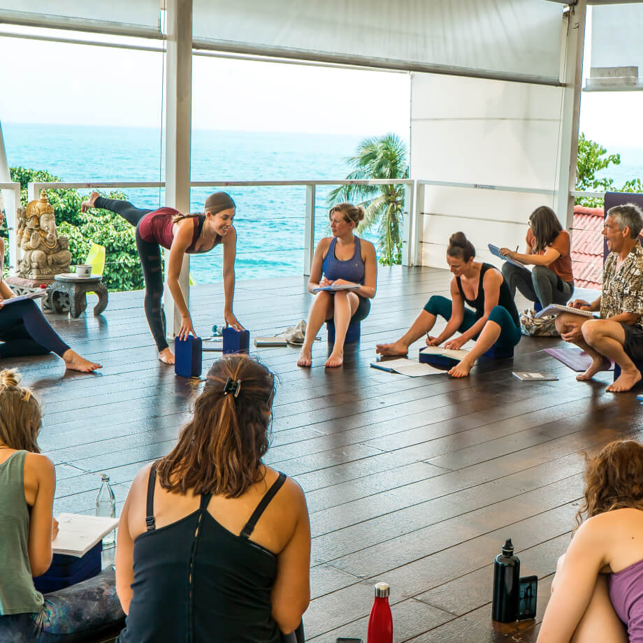 A circle of students watching a yoga teacher demonstrate a yoga pose in an open-air yoga studio overlooking crystal clear blue sea waters
