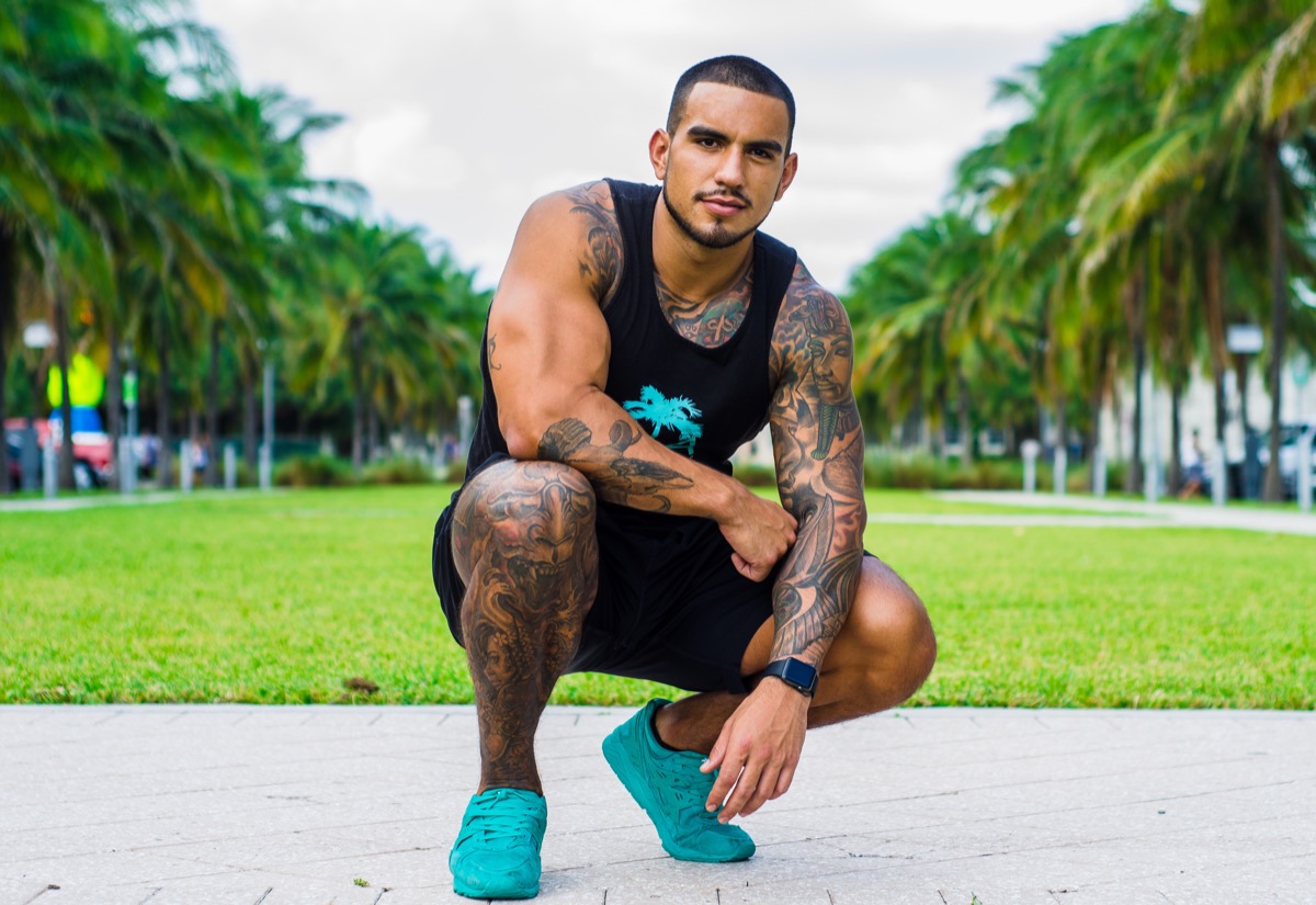 Heavily tattooed man in a black tank top and teal sneakers crouching on pavement in a Miami palm-tree-lined park