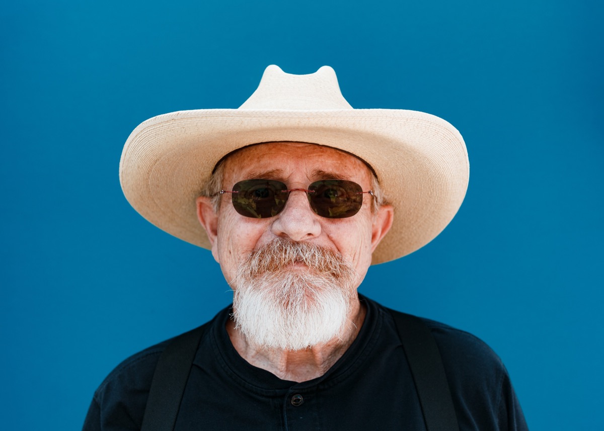 Portrait of an older man with white goatee and grey hair wearing a cream cowboy hat and dark sunglasses against a deep blue background