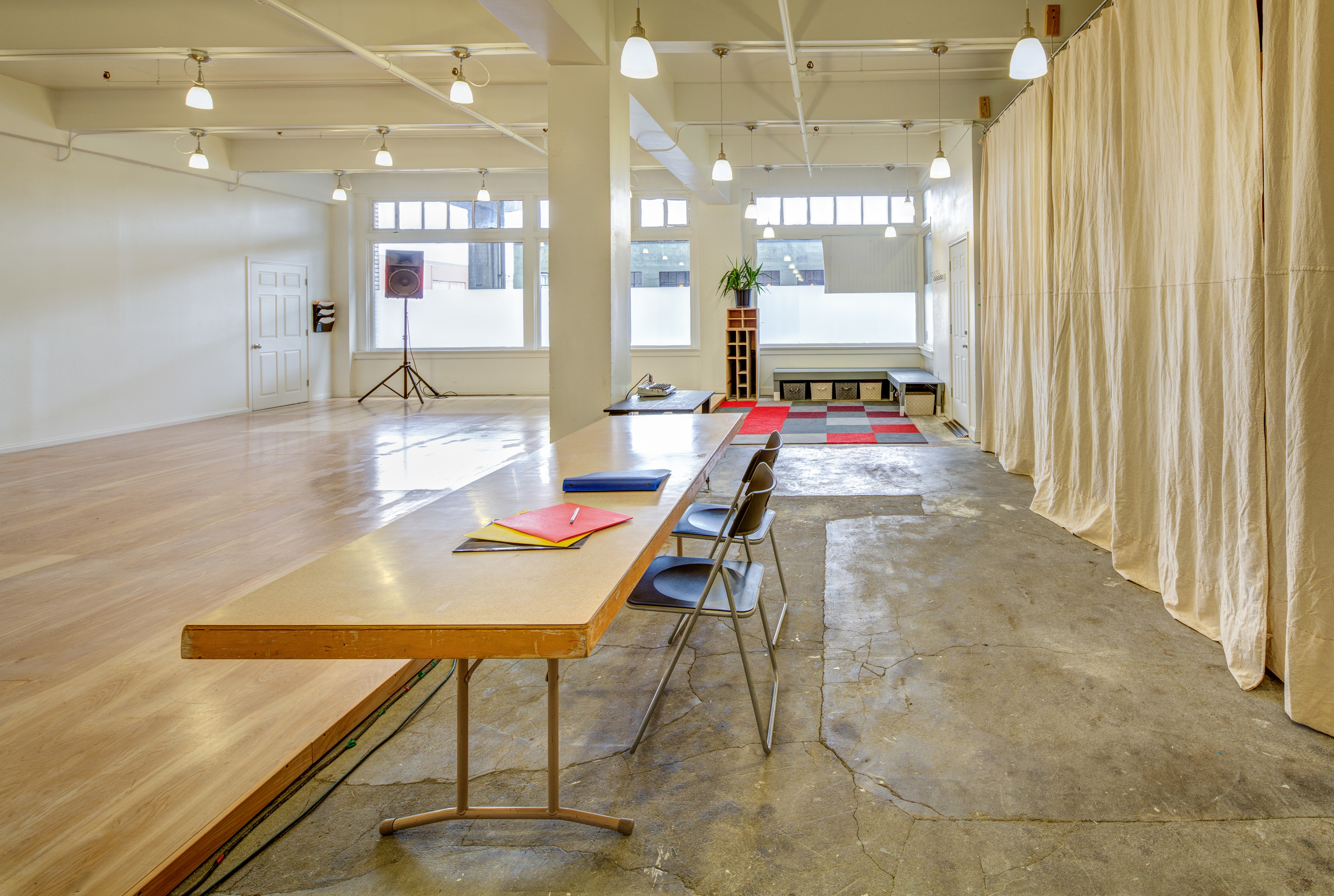 Bright photography studio with a long wooden table, folding chairs, hardwood and concrete flooring, beige curtains, and large windows