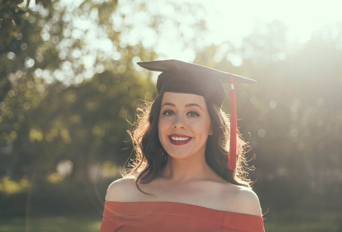 Smiling young woman wearing a graduation cap with a red tassel and an off-shoulder red top, photographed outdoors at golden hour