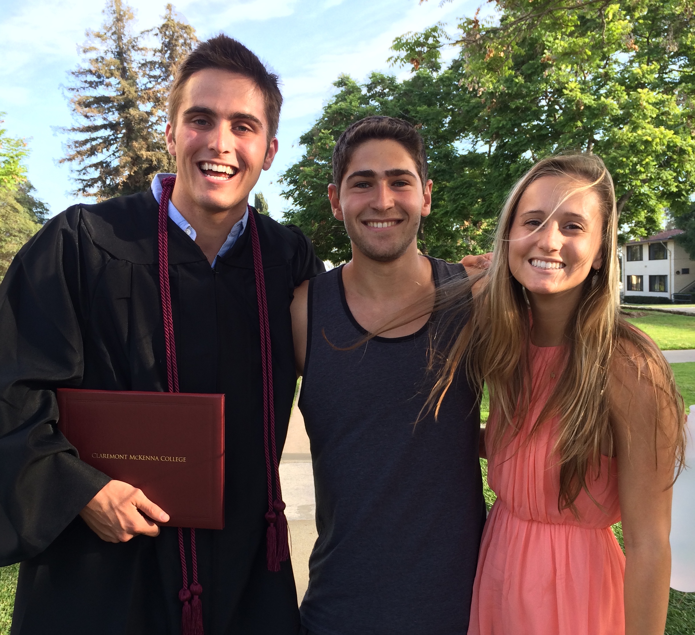 Three young friends posing at graduation, with the male graduate in a black gown holding a Claremont McKenna College diploma