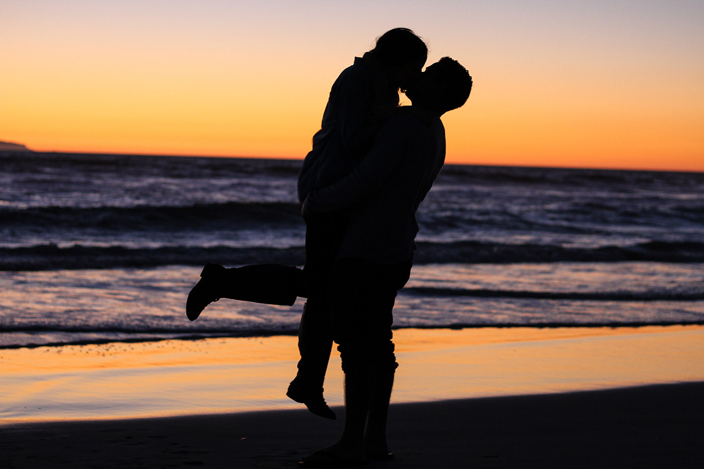 Silhouette of an engaged couple kissing on the beach at sunset, with the man lifting the woman in his arms over the ocean