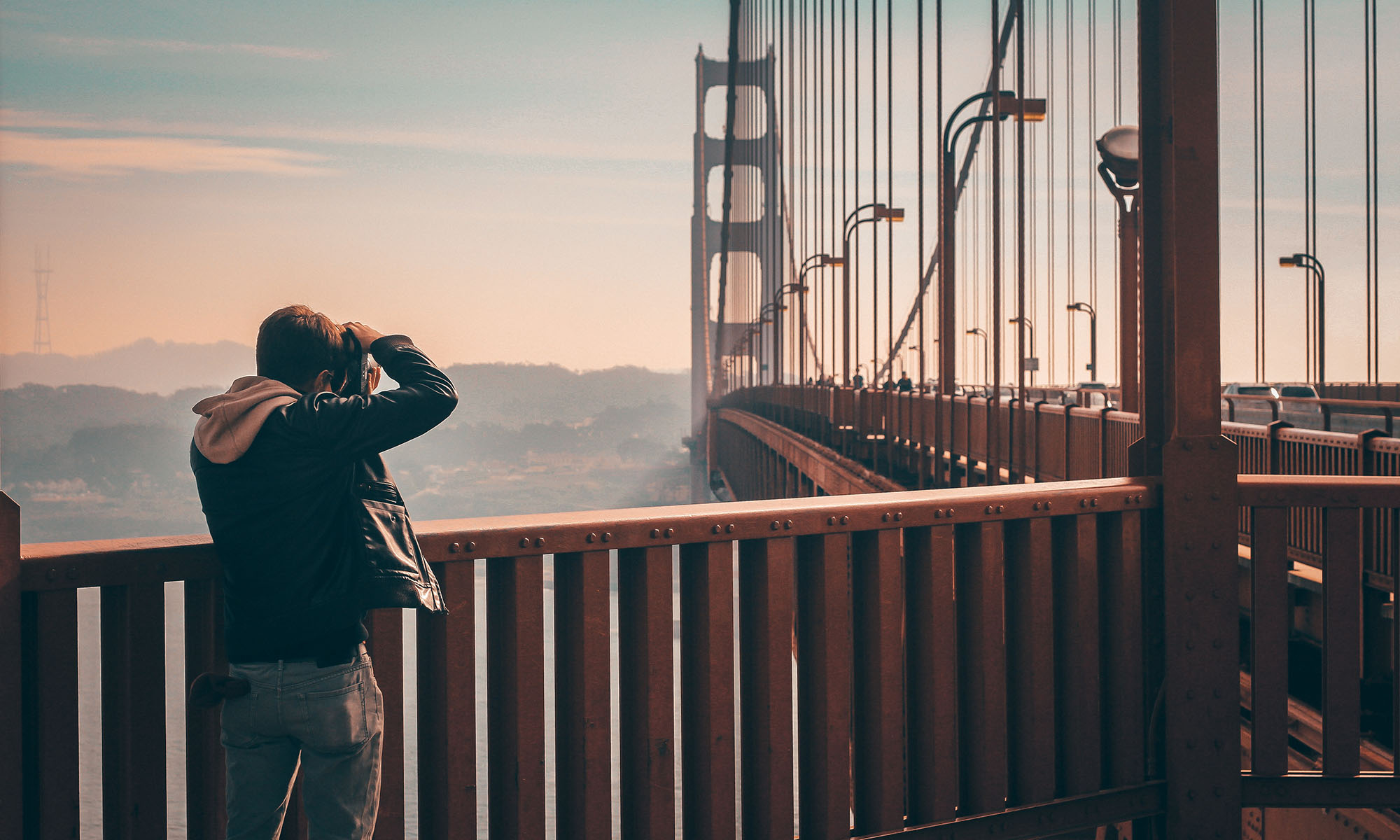 Photographer with hood standing on the Golden Gate Bridge walkway, taking a photo of the bridge towers at sunset