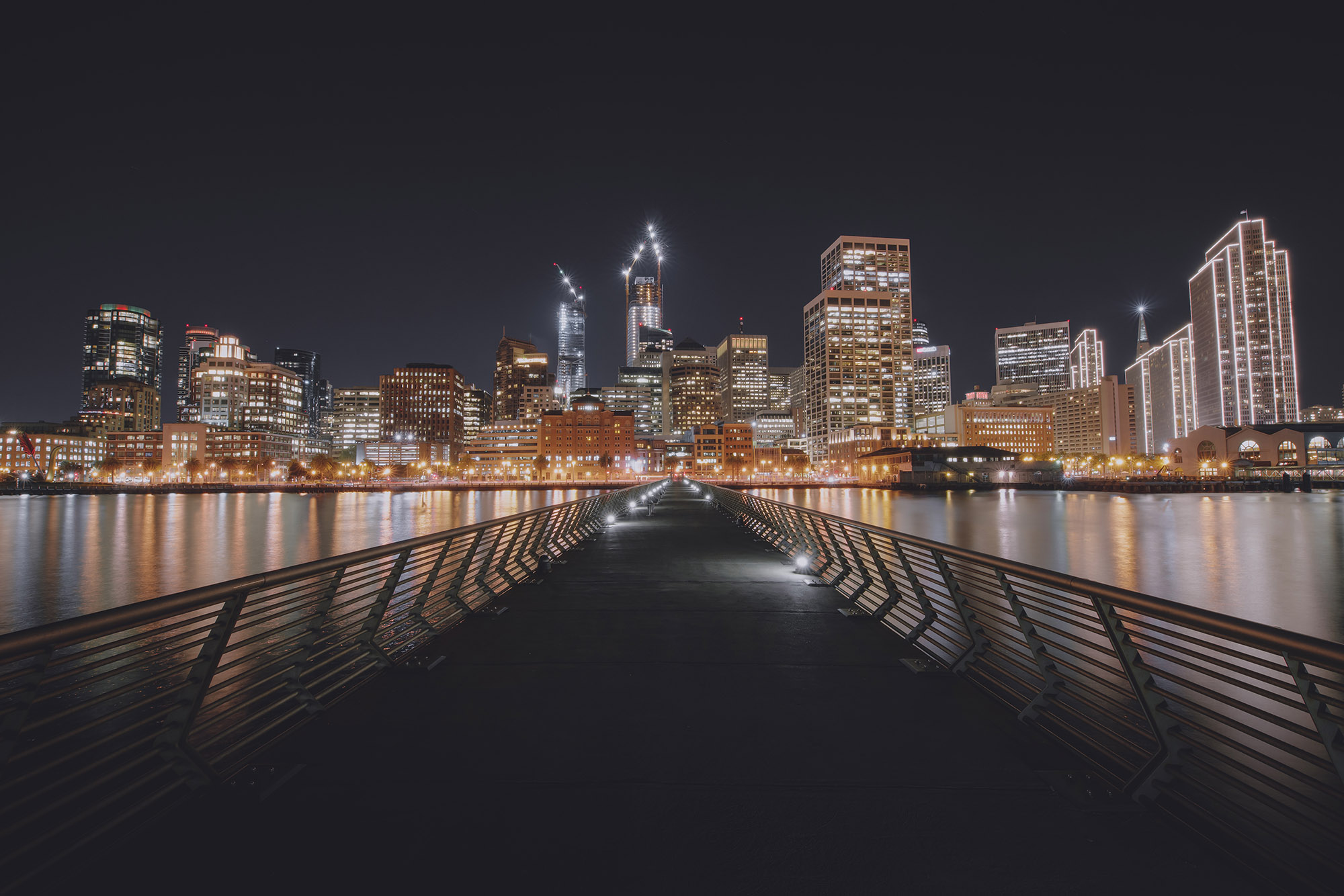 Pier 14 wooden walkway leading toward the illuminated San Francisco downtown skyline at night reflecting on the bay