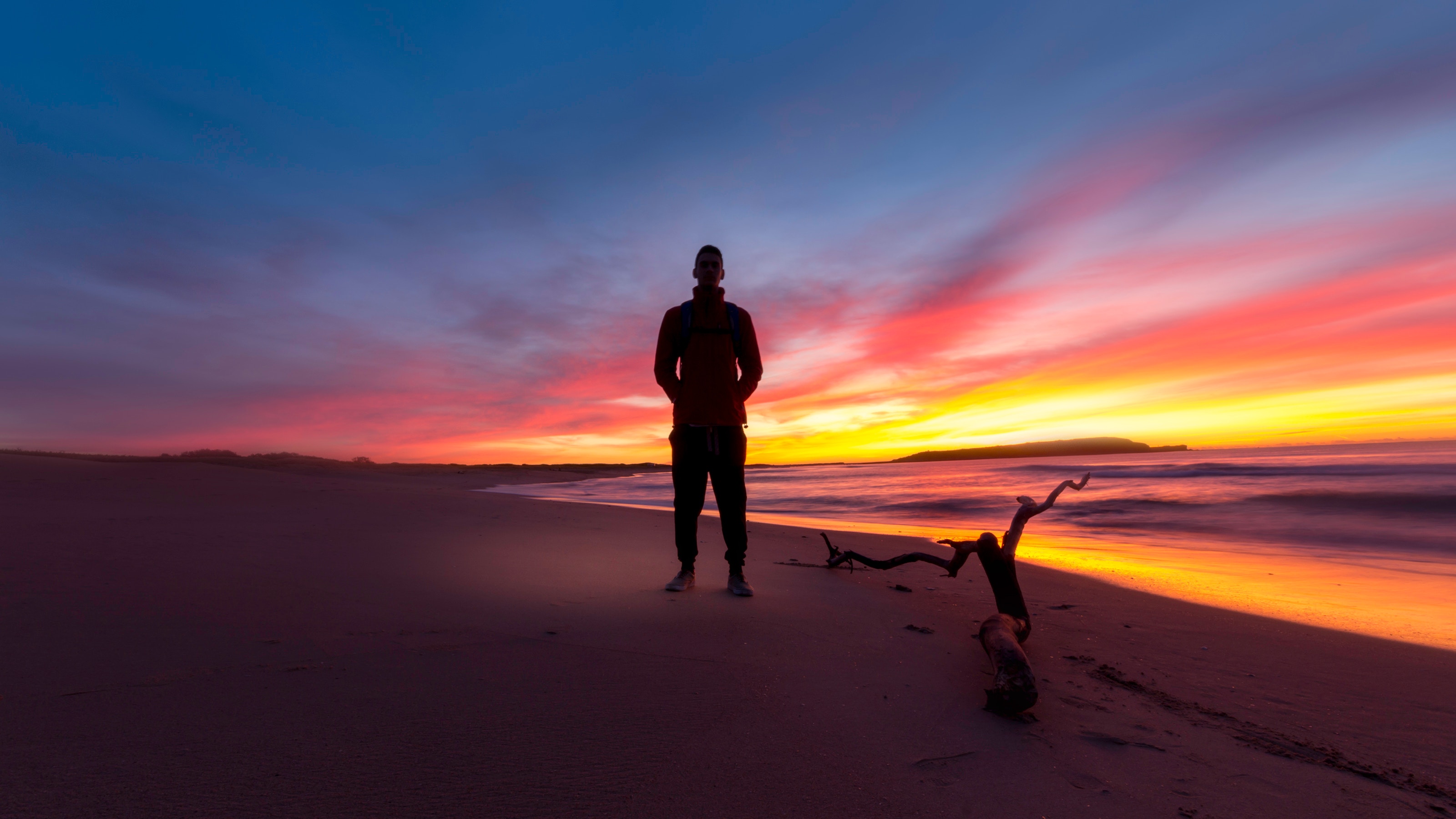 Silhouette of a man with a backpack standing on a sandy beach at sunset with vivid pink and orange sky and driftwood beside him