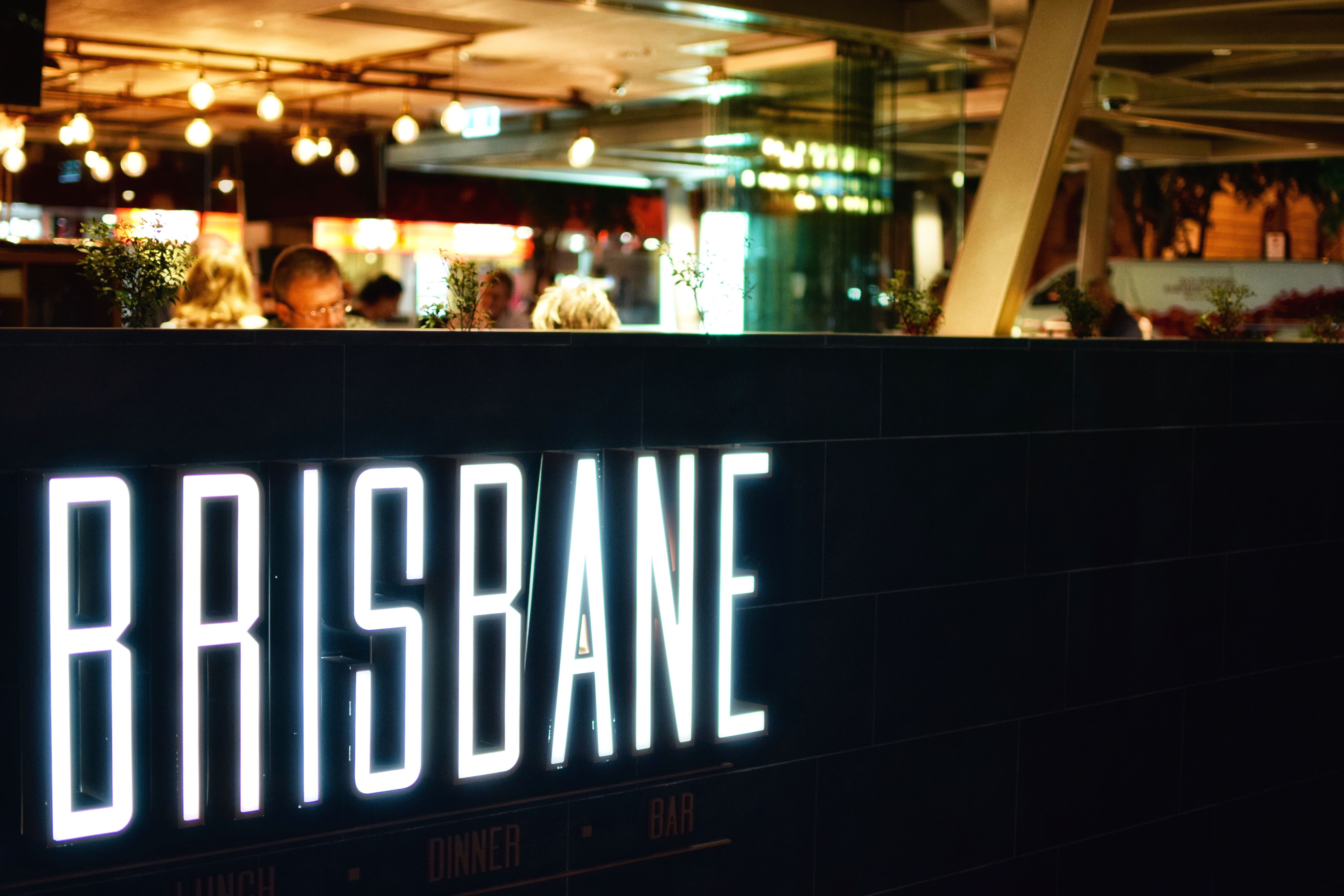 Illuminated white BRISBANE neon sign on a dark tiled wall inside a restaurant or bar with diners visible behind