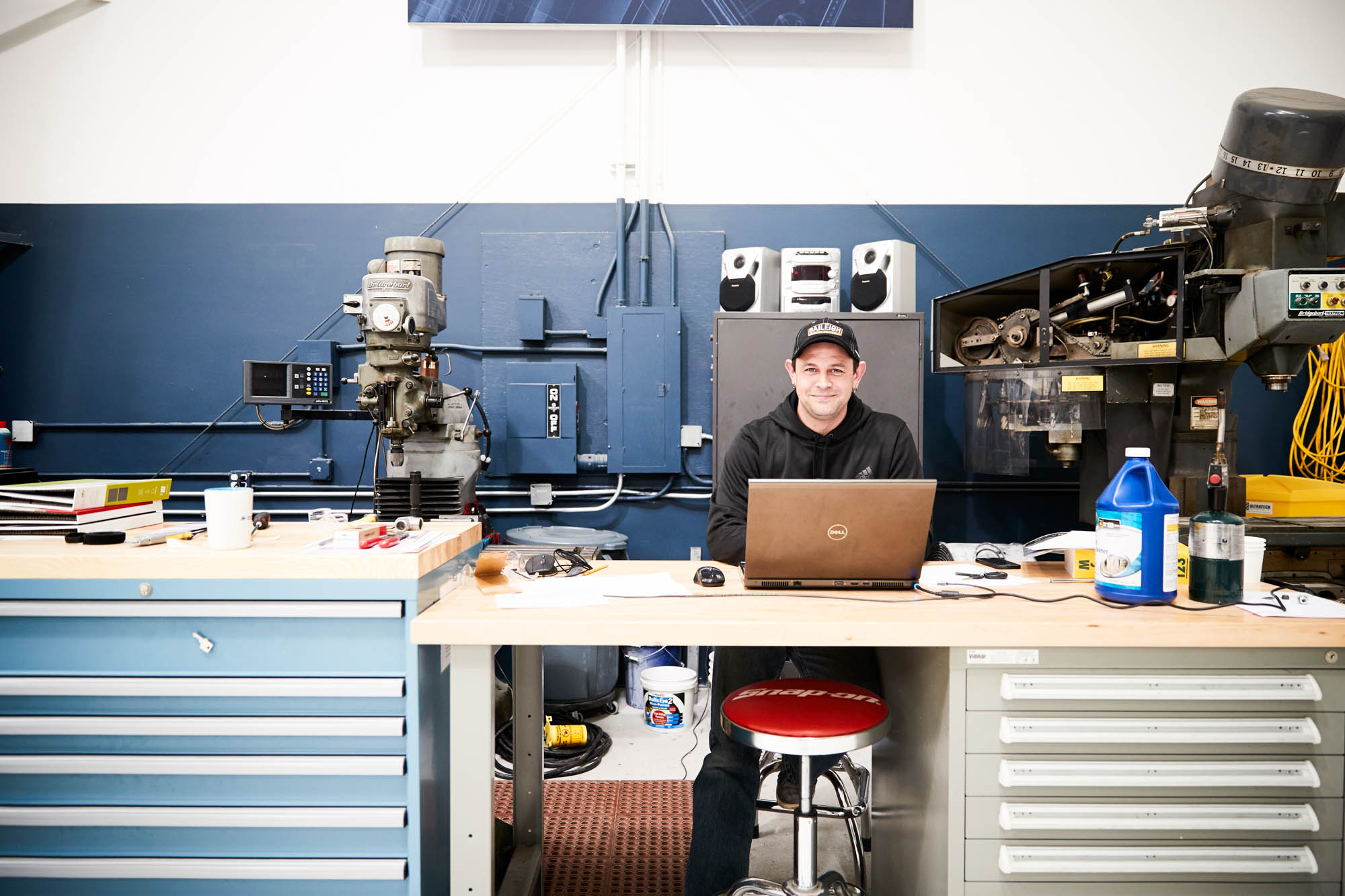 Boom Supersonic technician seated at a workshop desk with a Dell laptop, surrounded by milling machines and metalworking equipment