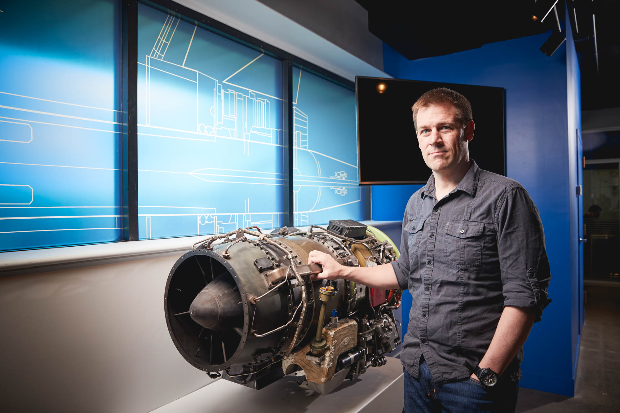 Boom Supersonic engineer posing next to a dismounted jet engine inside the office with engineering schematics on a backlit blue wall
