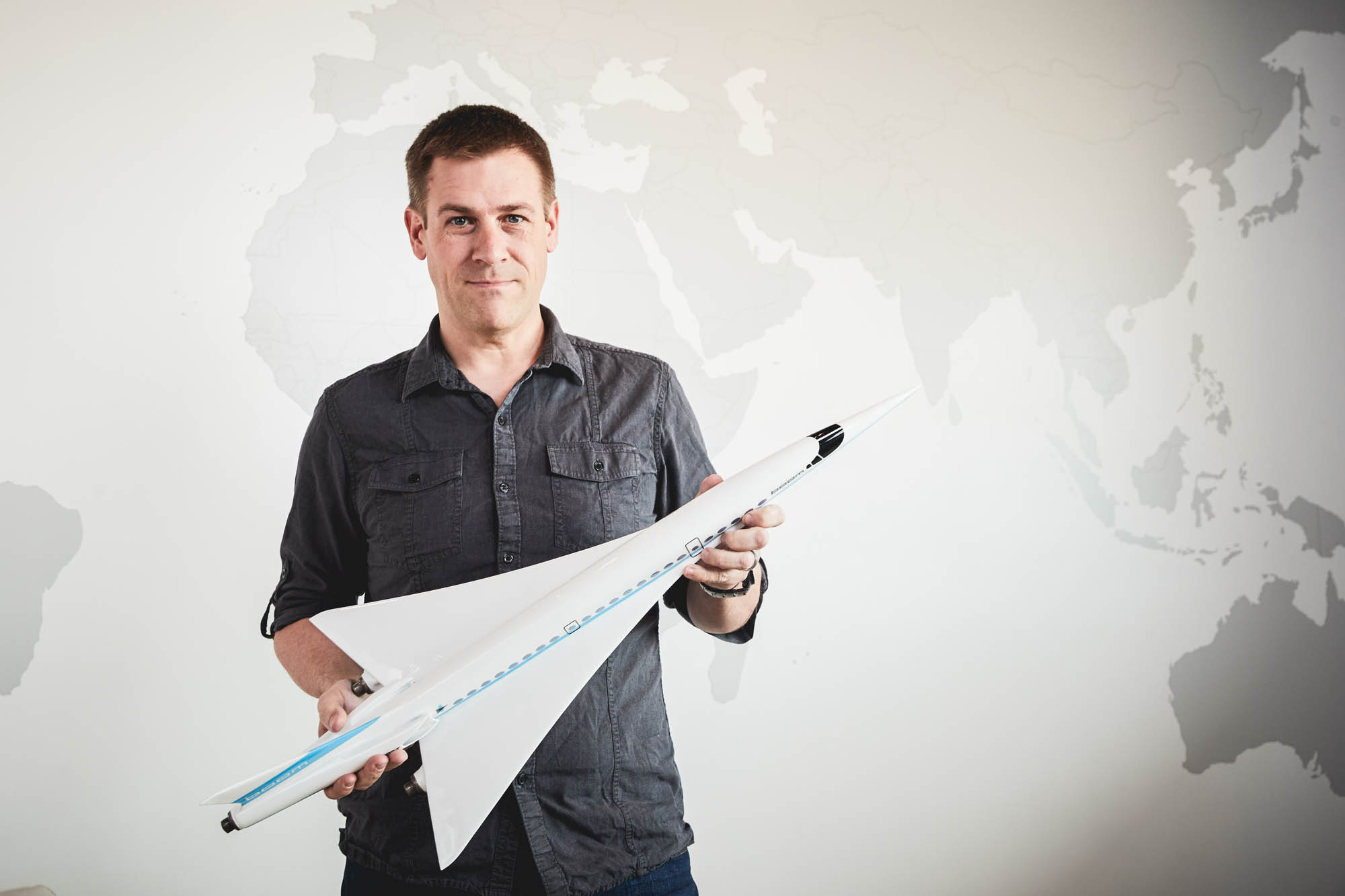 Boom Supersonic team member in a dark shirt holding a model of the Overture supersonic airliner in front of a world map wall